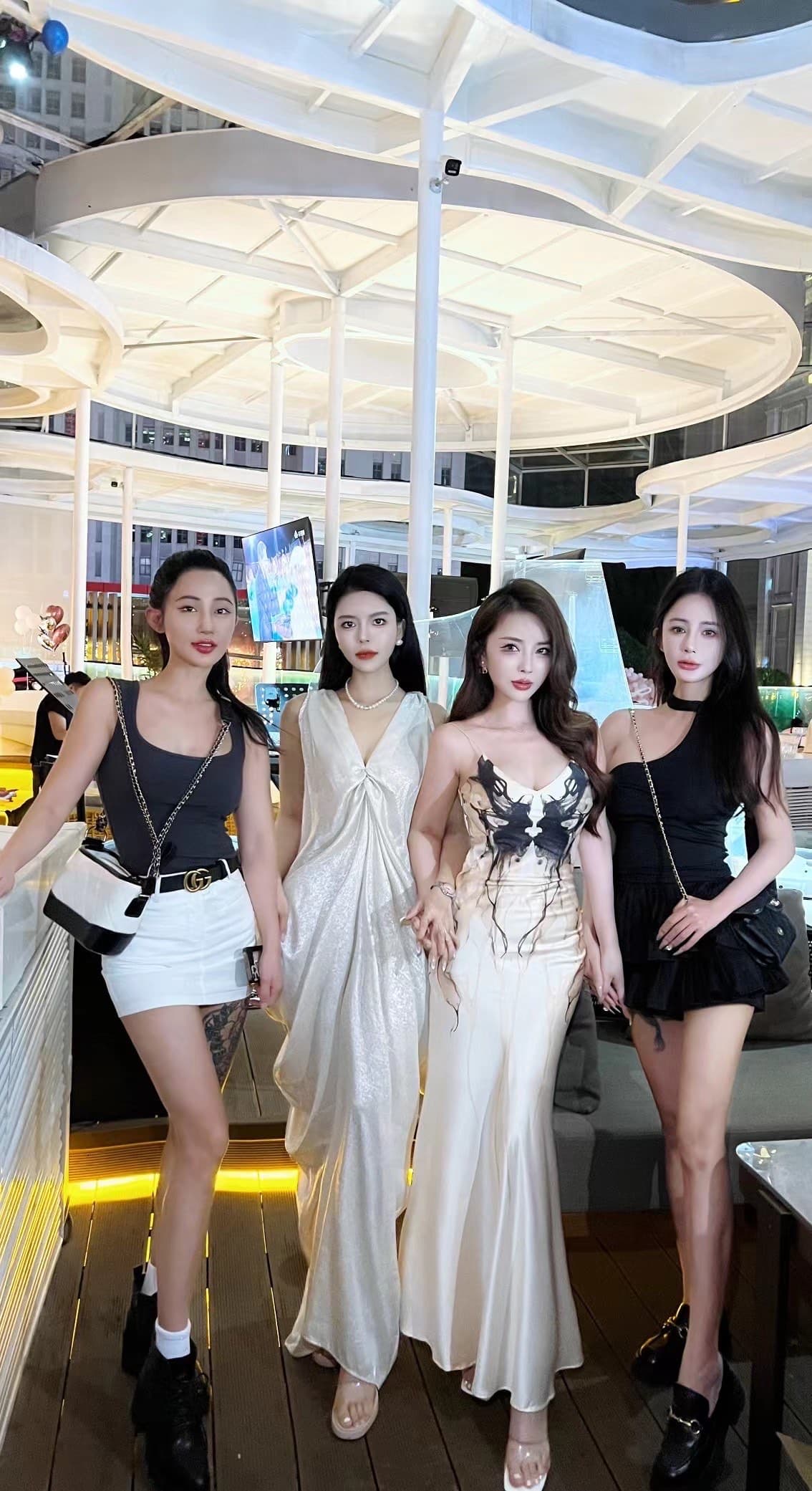 Four stylish women in black and white outfits pose at a modern rooftop lounge.