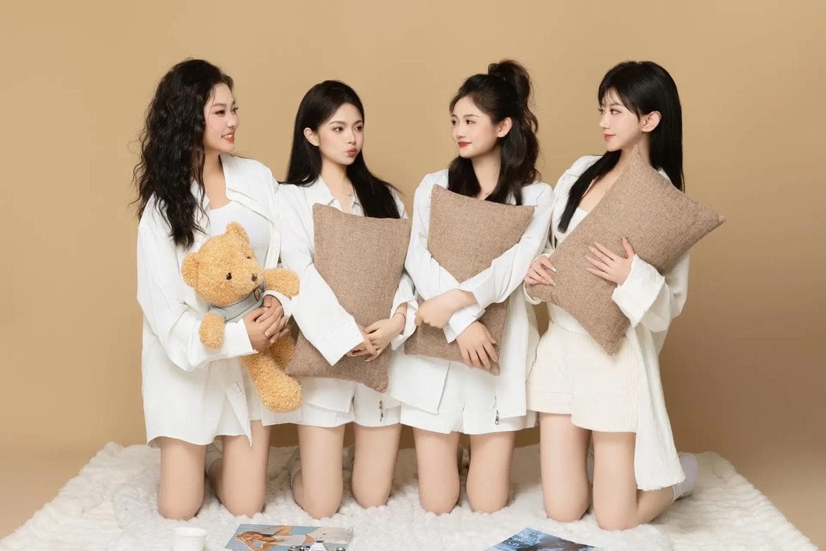 Four women in white shirts kneel on a rug, holding pillows and a teddy bear.